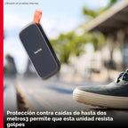 SSD portátil SanDisk 1TB en caída libre al aire libre junto a una zapatilla, texto: protección contra caídas de hasta dos metros permite que esta unidad resista golpes
